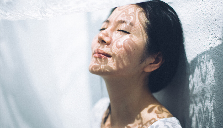 A woman stands with her back against a wall, eyes closed and head tilted towards the light filtering through a fabric hung overhead, casting a lace pattern on her face.