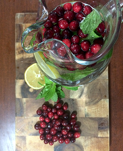 The top view of a pitcher of cranberry and mint infused water surrounded by cranberries, a lemon slice, and mint leaves arranged on a cutting board