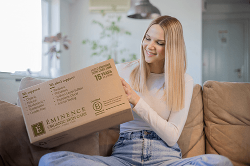 A woman holding an Eminence Organics box while sitting on a couch
