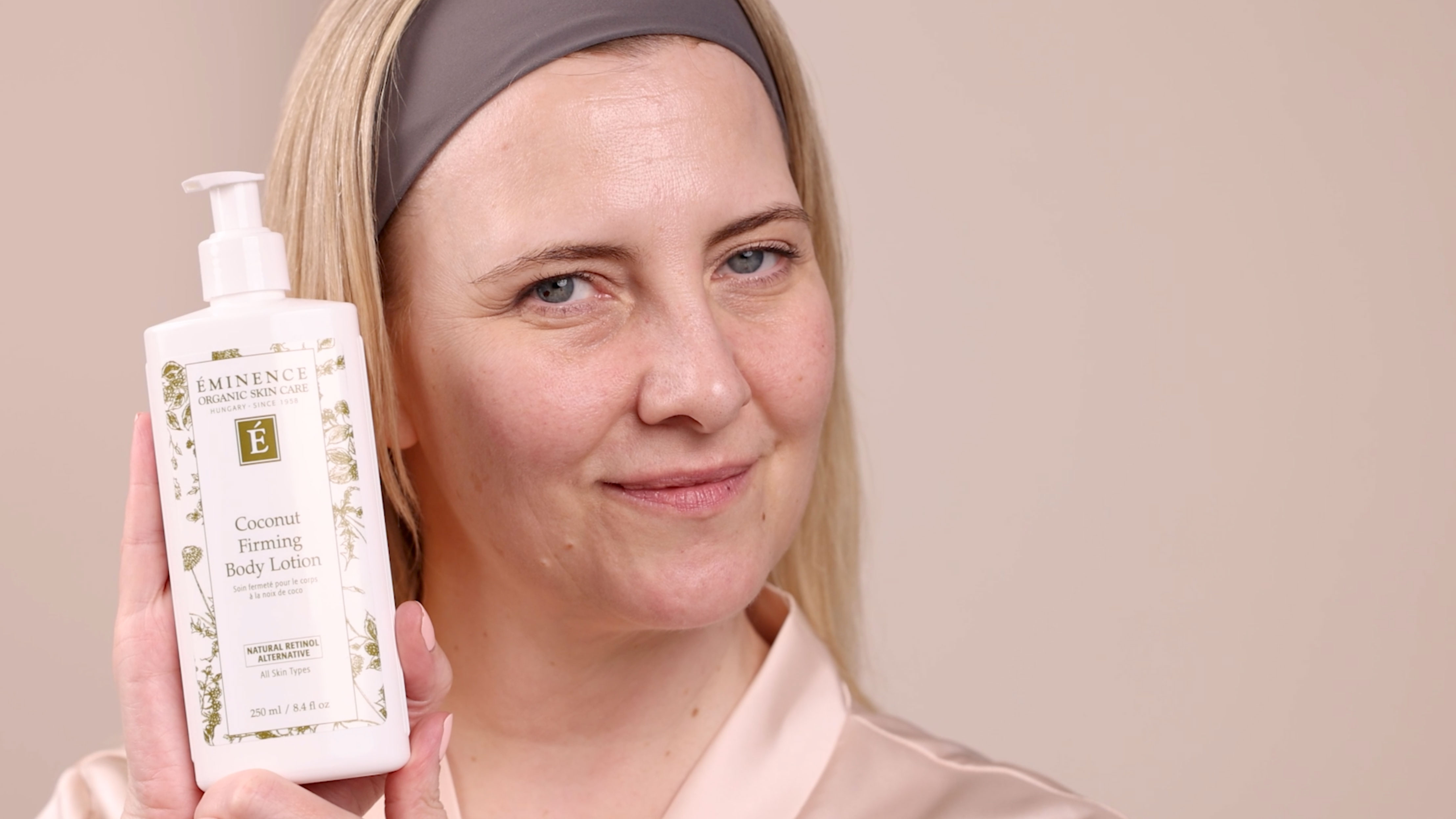 A woman holding the Eminence Organics Coconut Firming Body Lotion bottle beside her face