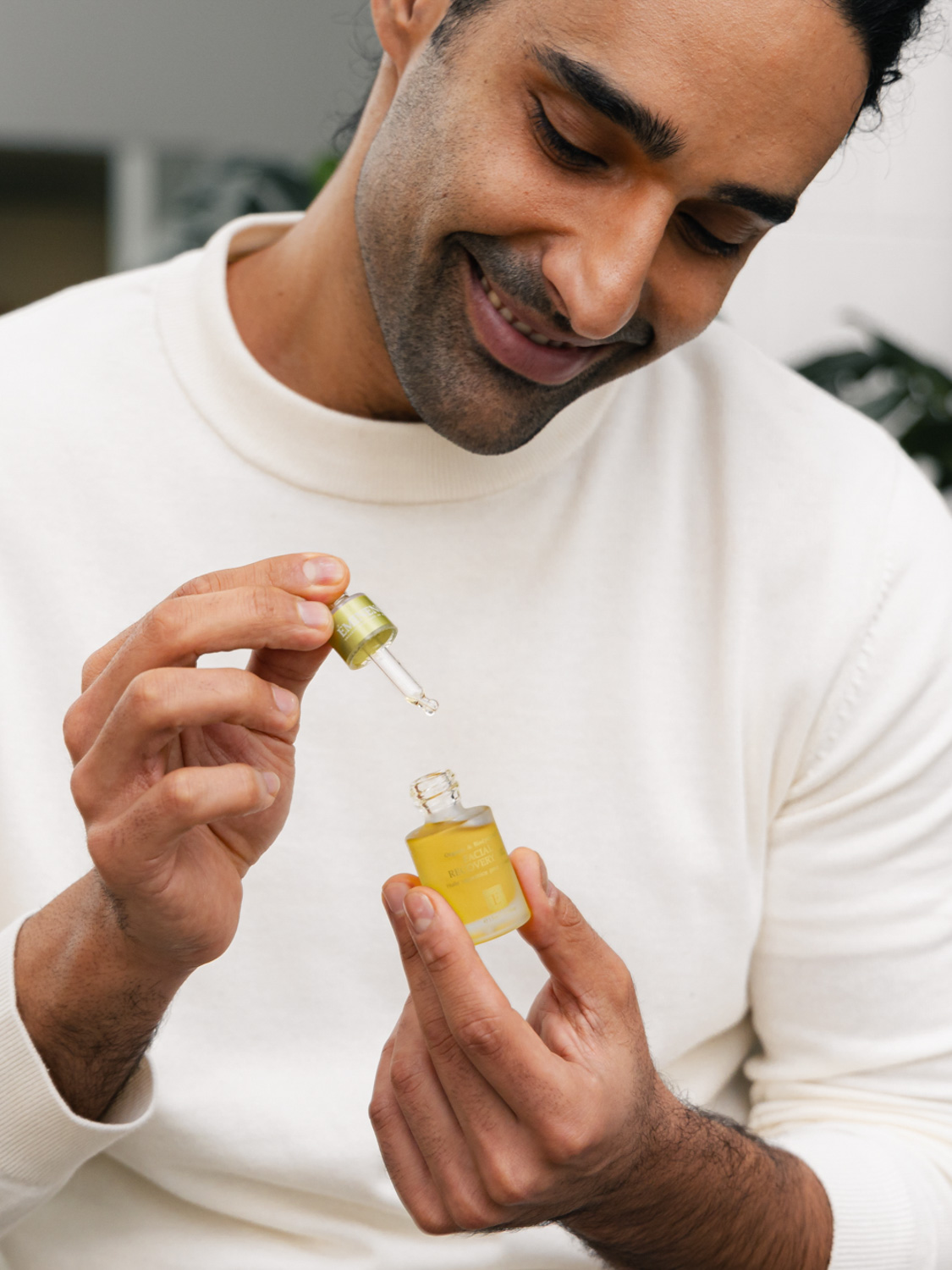 A man holding the Eminence Organics Facial Recovery Oil glass bottle in one hand and the dropper in the other hand