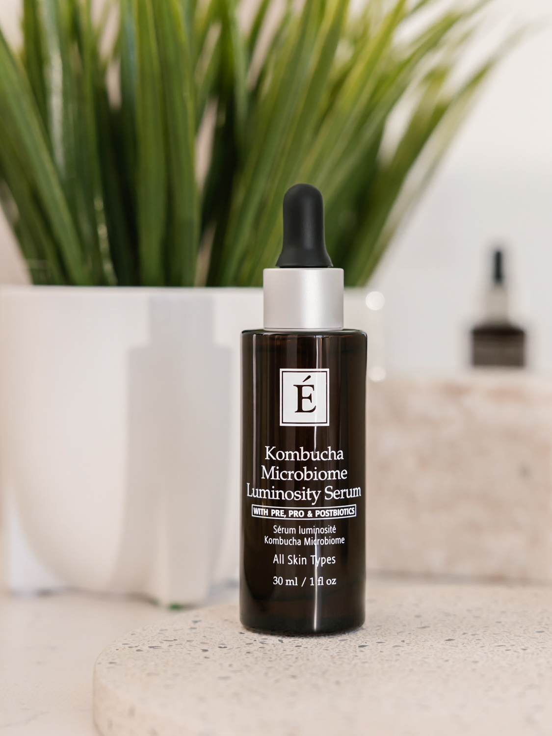 Eminence Organics Kombucha Microbiome Luminosity Serum bottle in front of a fern plant