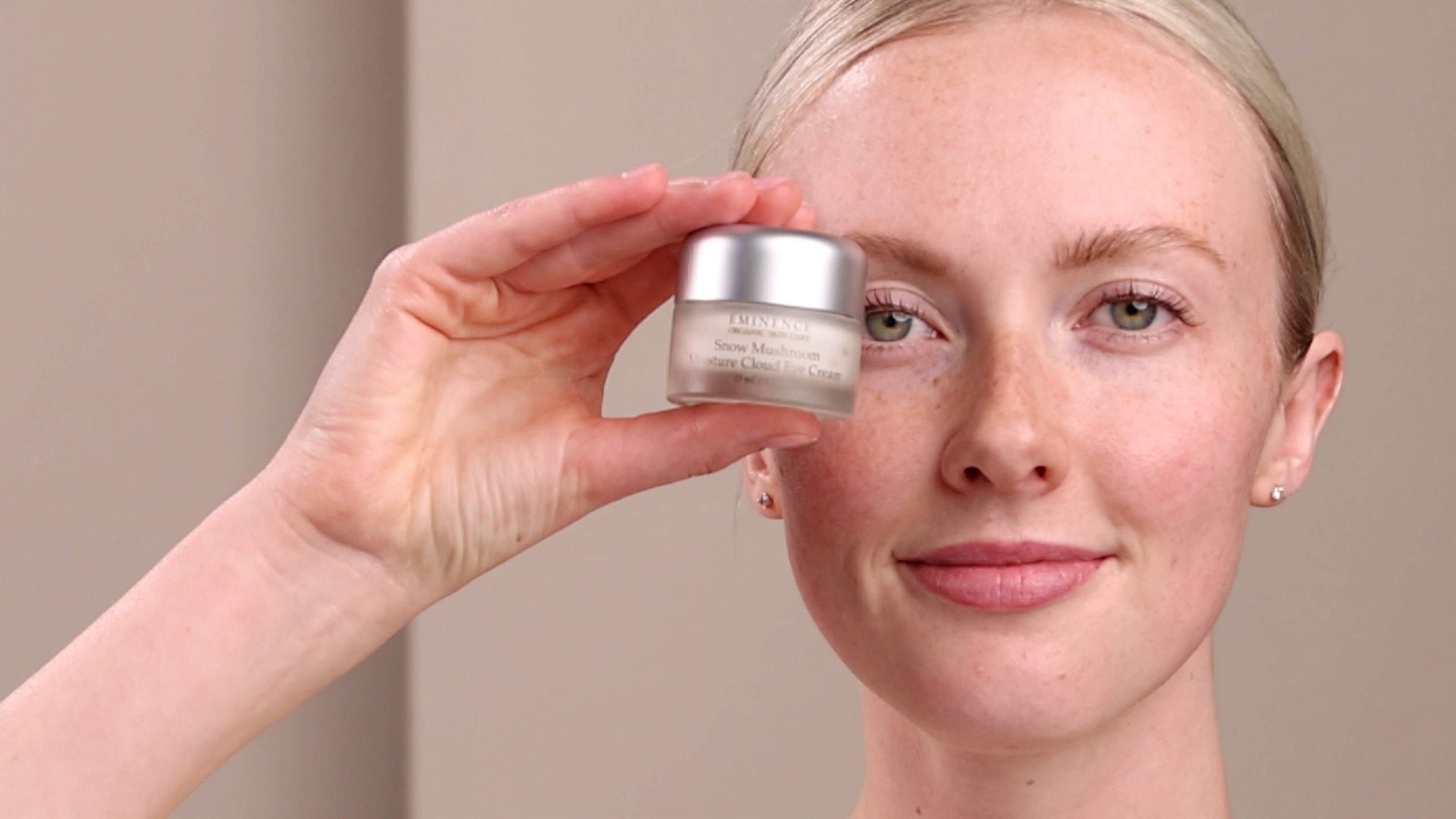 A woman holding an open jar of the Eminence Organics Snow Mushroom Moisture Cloud Eye Cream and showing her hand with a swatch of the eye cream