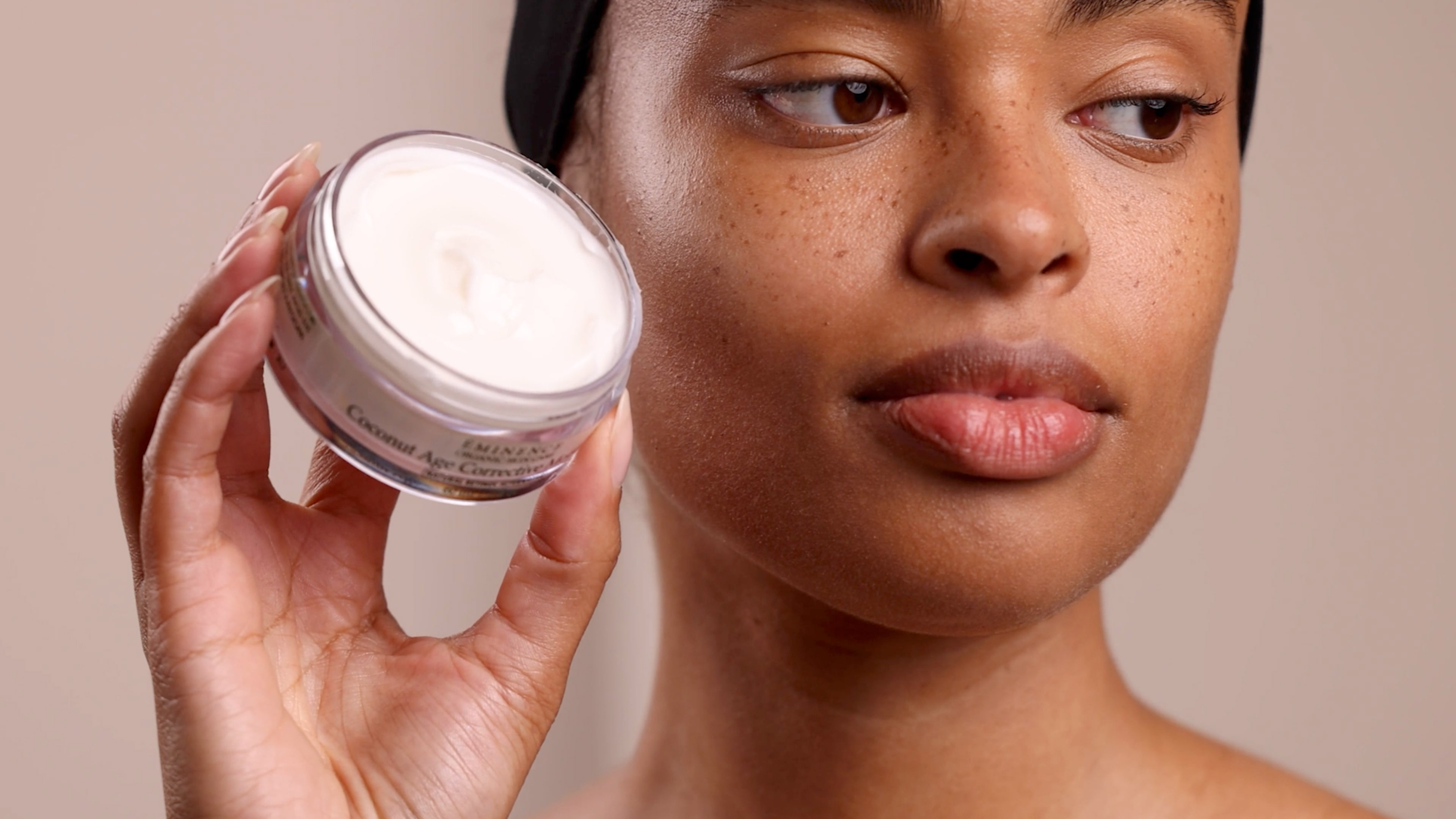 Woman holding up Eminence Organics Coconut Age Corrective Moisturizer next to face