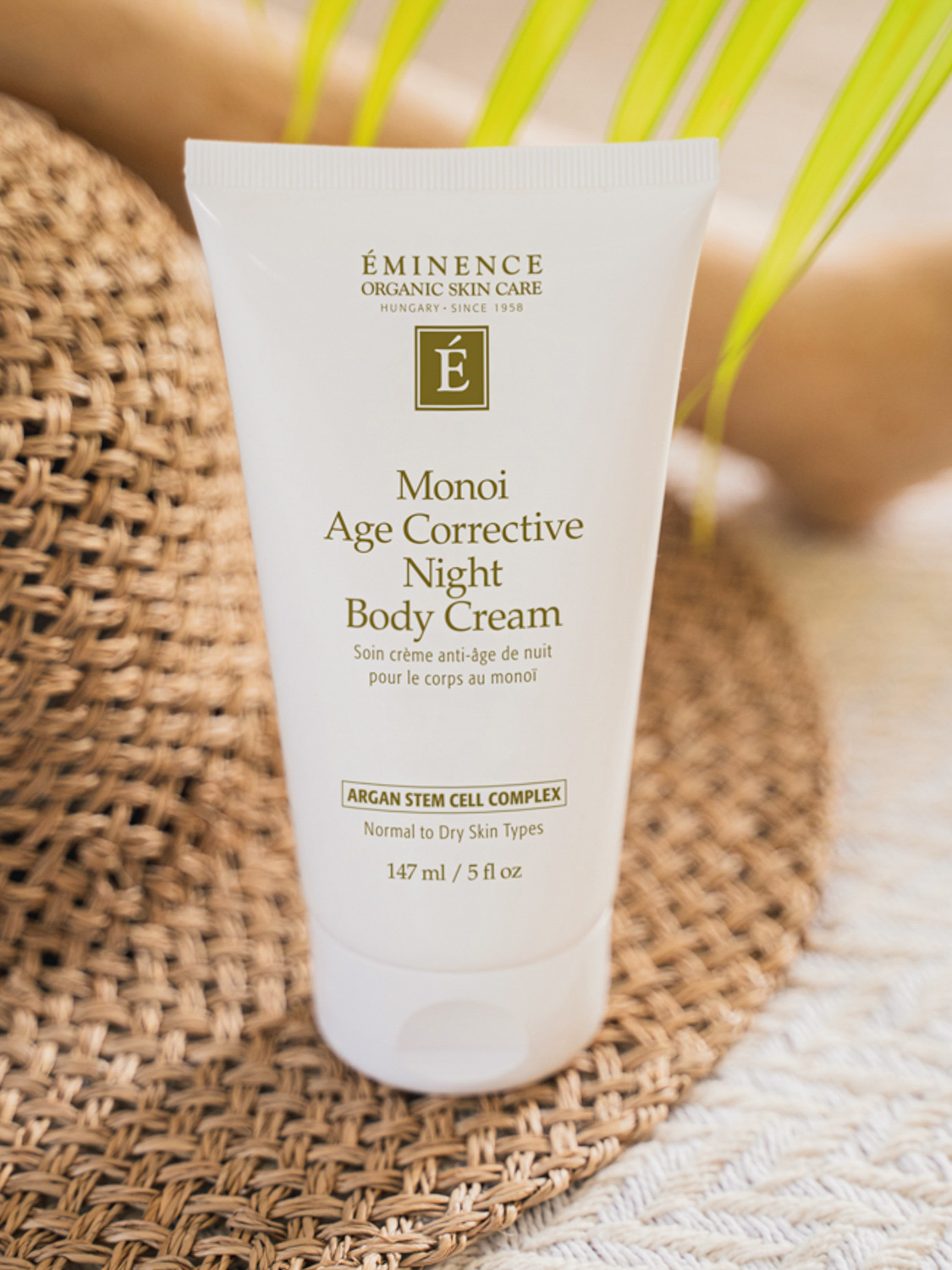 The Eminence Organics Monoi Age Corrective Night Body Cream tube beside a sun hat