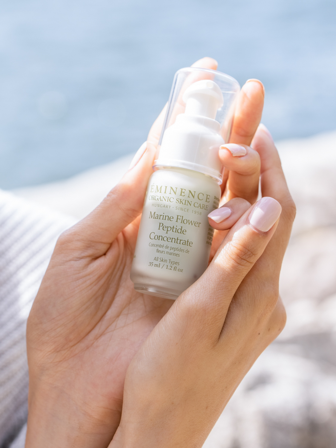 A hand holding the Eminence Organics Marine Flower Peptide Concentrate glass bottle next to the ocean