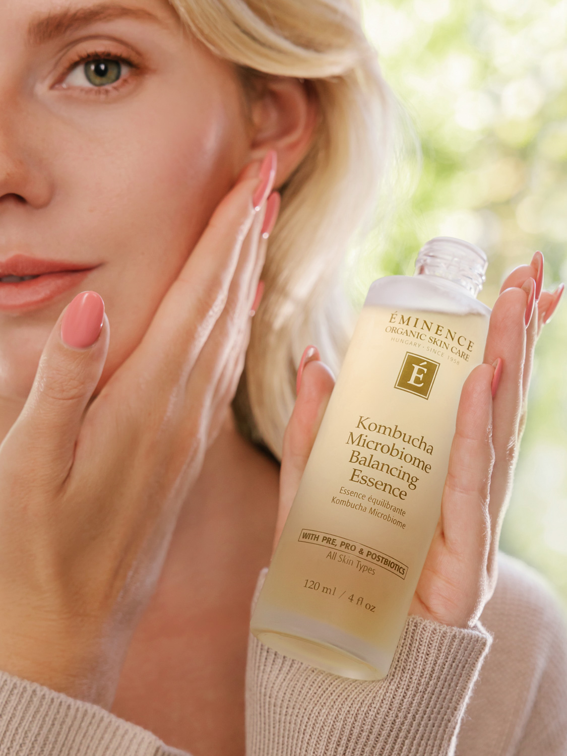 A woman holding the Eminence Organics Kombucha Microbiome Balancing Essence glass bottle and applying the essence onto her face