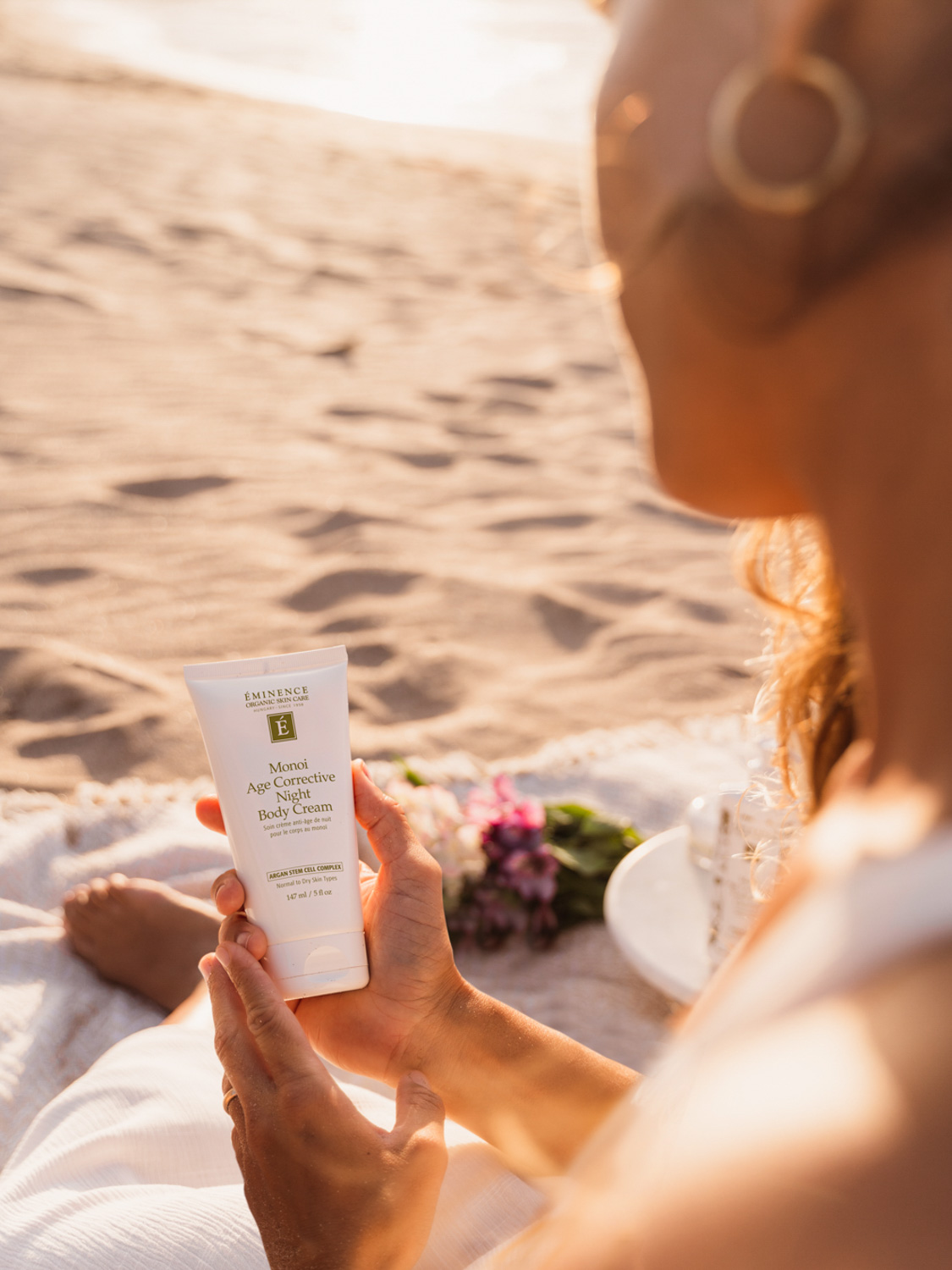 A woman holding the Eminence Organics Monoi Age Corrective Night Body Cream on the beach