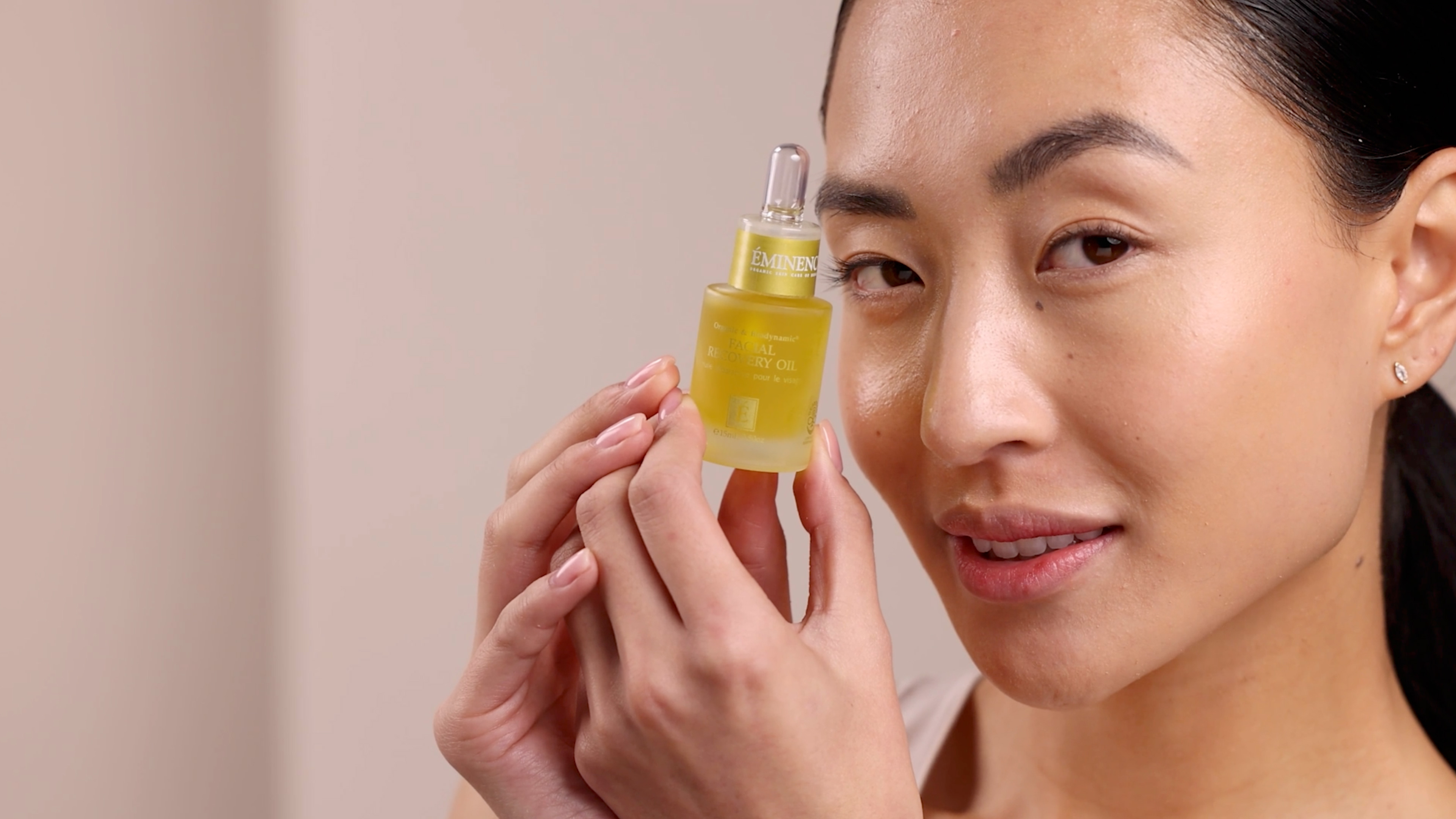 A woman holding the Eminence Organics Facial Recovery Oil glass bottle next to her face