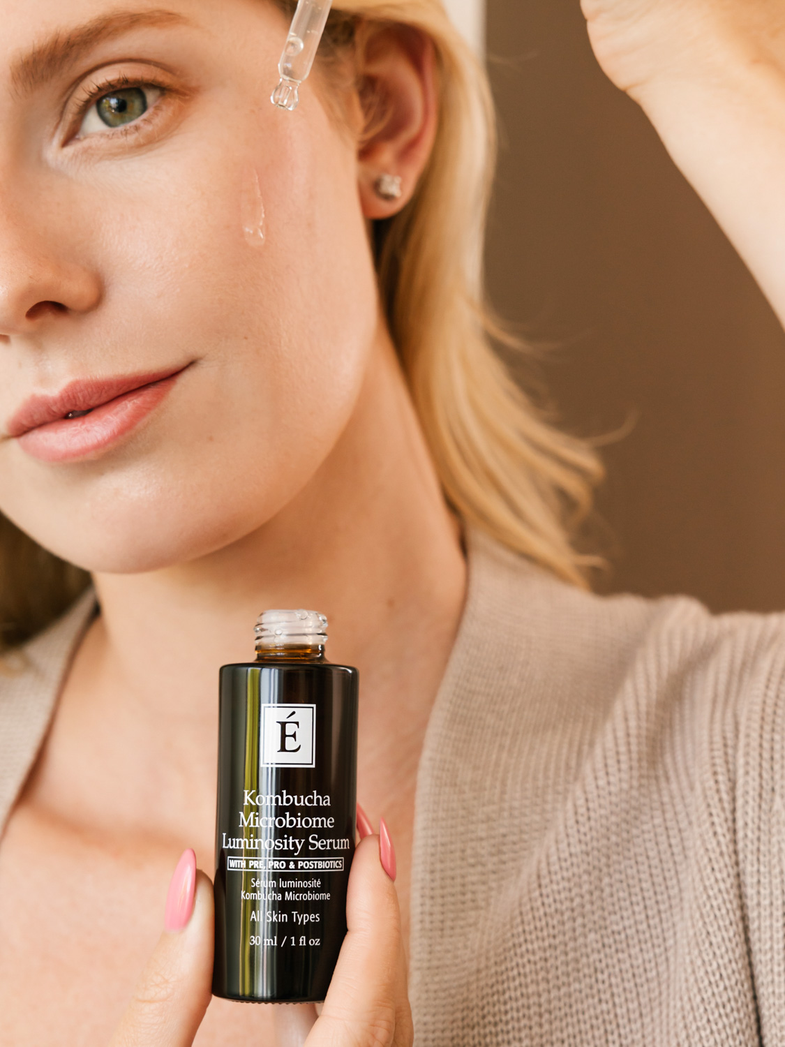 A woman holding the Eminence Organics Kombucha Microbiome Luminosity Serum dropper to her face with one hand and the serum bottle with the other hand