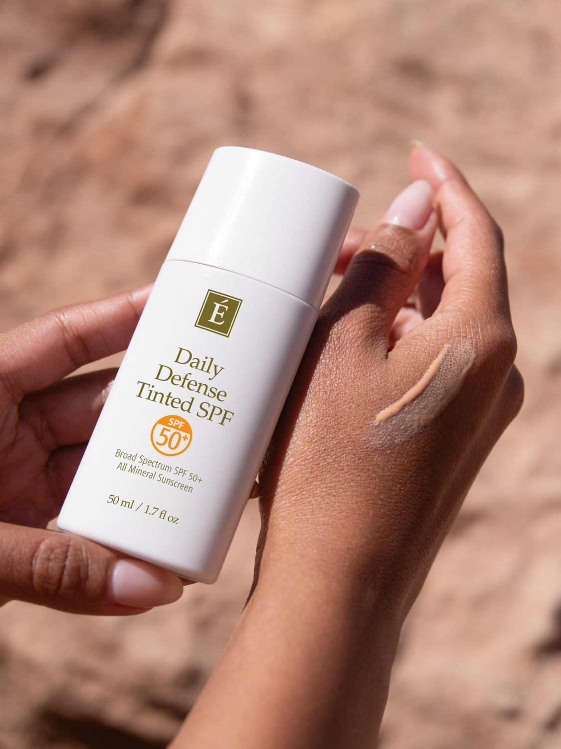 A woman holding the Eminence Organics Daily Defense Tinted SPF and a swatch of the SPF on her other hand