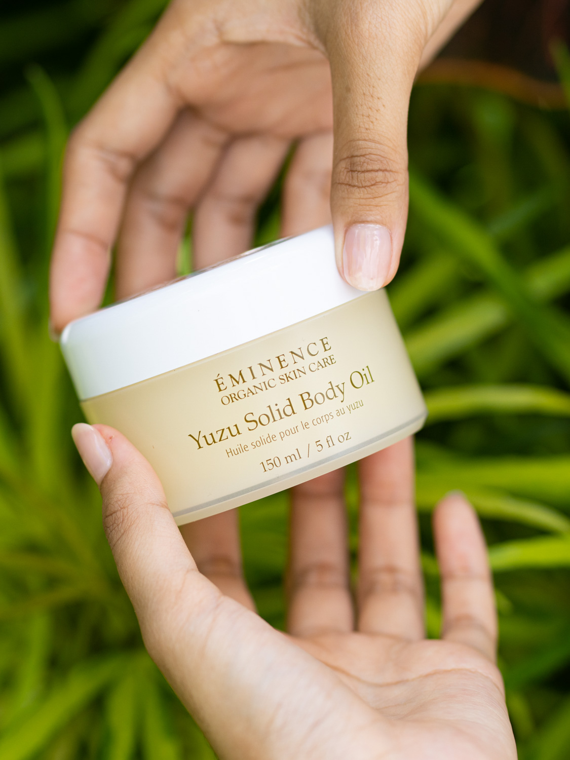 Hands holding the Eminence Organics Yuzu Solid Body Oil with a background of green leaves