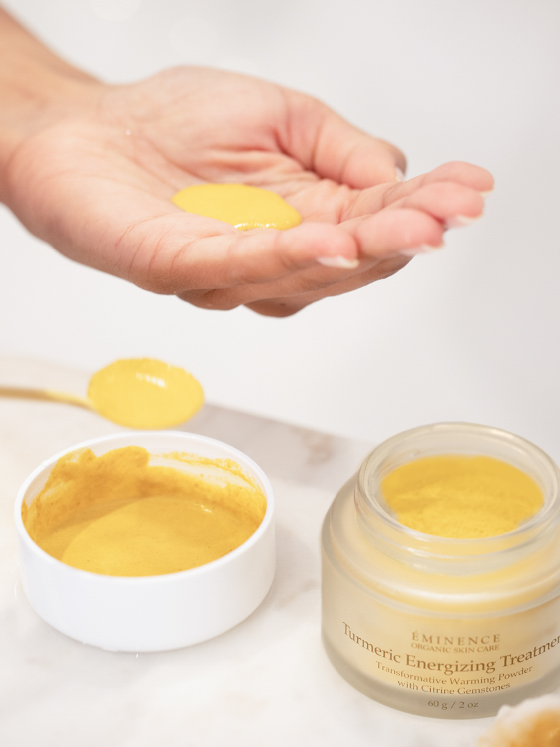 A hand with an amount of the Eminence Organics Turmeric Energizing Treatment and the open jar on the background