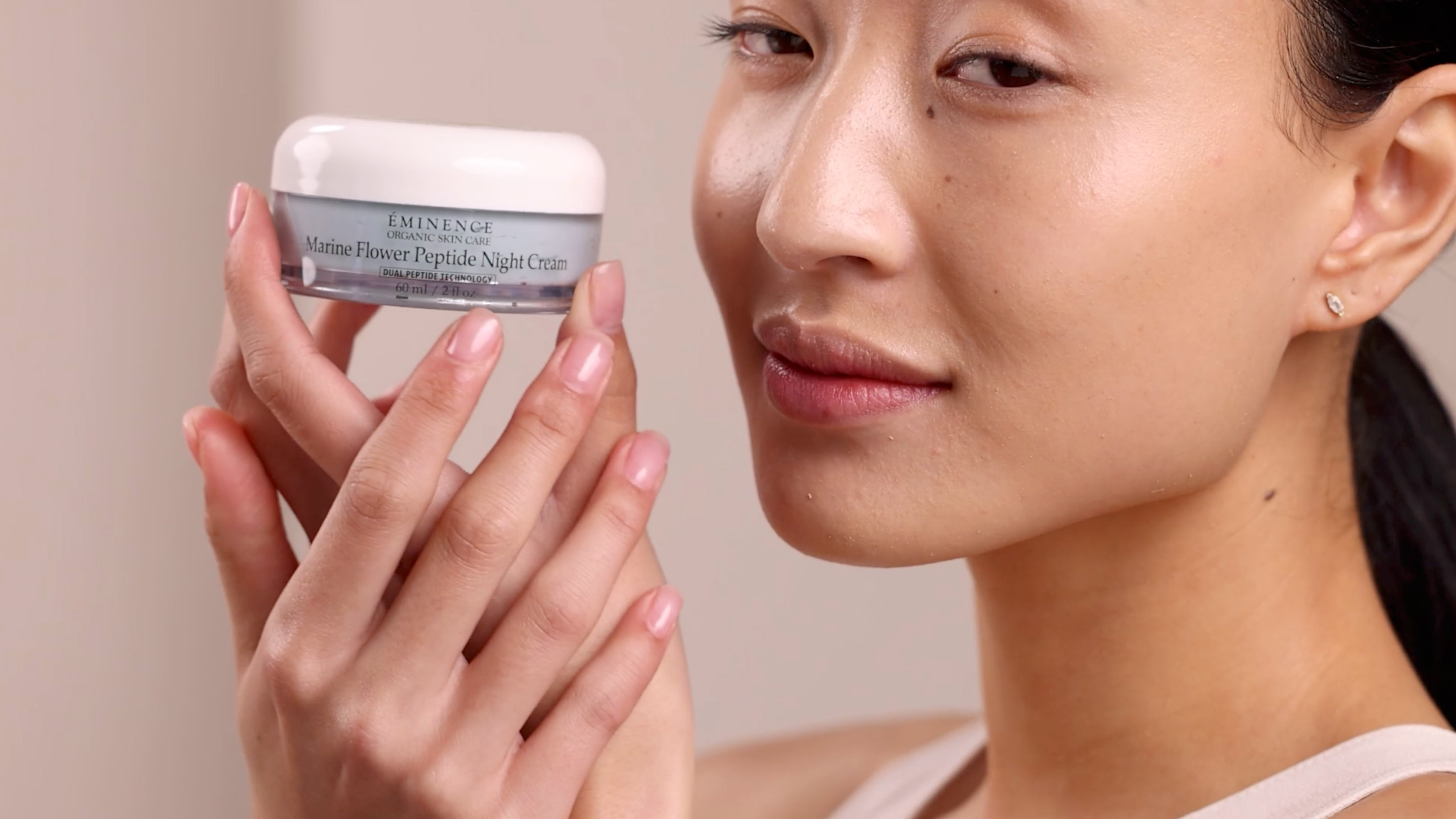 A woman holding the Eminence Organics Marine Flower Peptide Night Cream jar next to her face