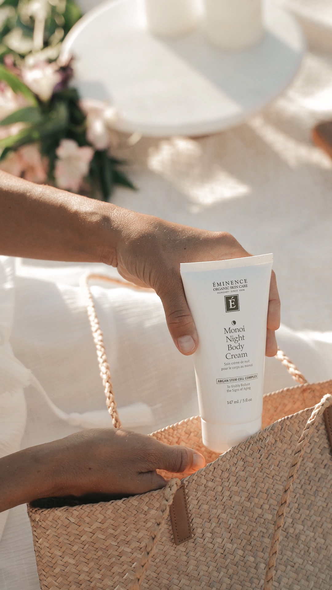 A hand placing the Eminence Organics Monoi Age Corrective Night Body Cream into a large purse