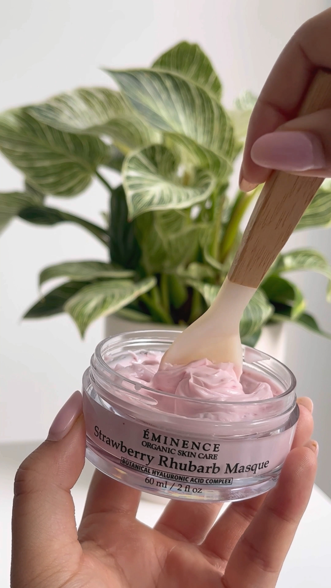 A hand holding an open jar of the Eminence Organics Strawberry Rhubarb Masque with a spoon on it