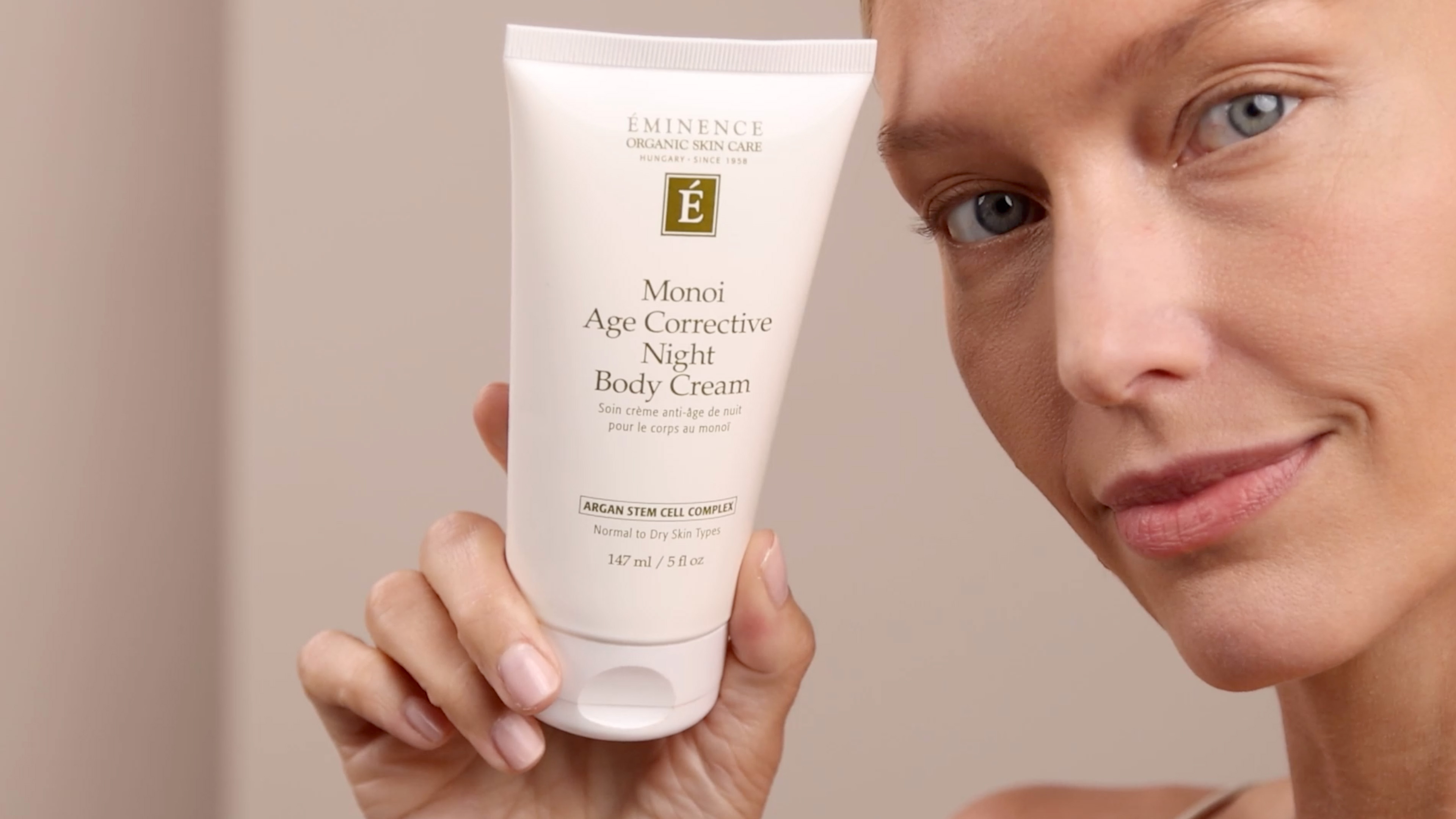 A woman holding the Eminence Organics Monoi Age Corrective Night Body Cream tube beside her face