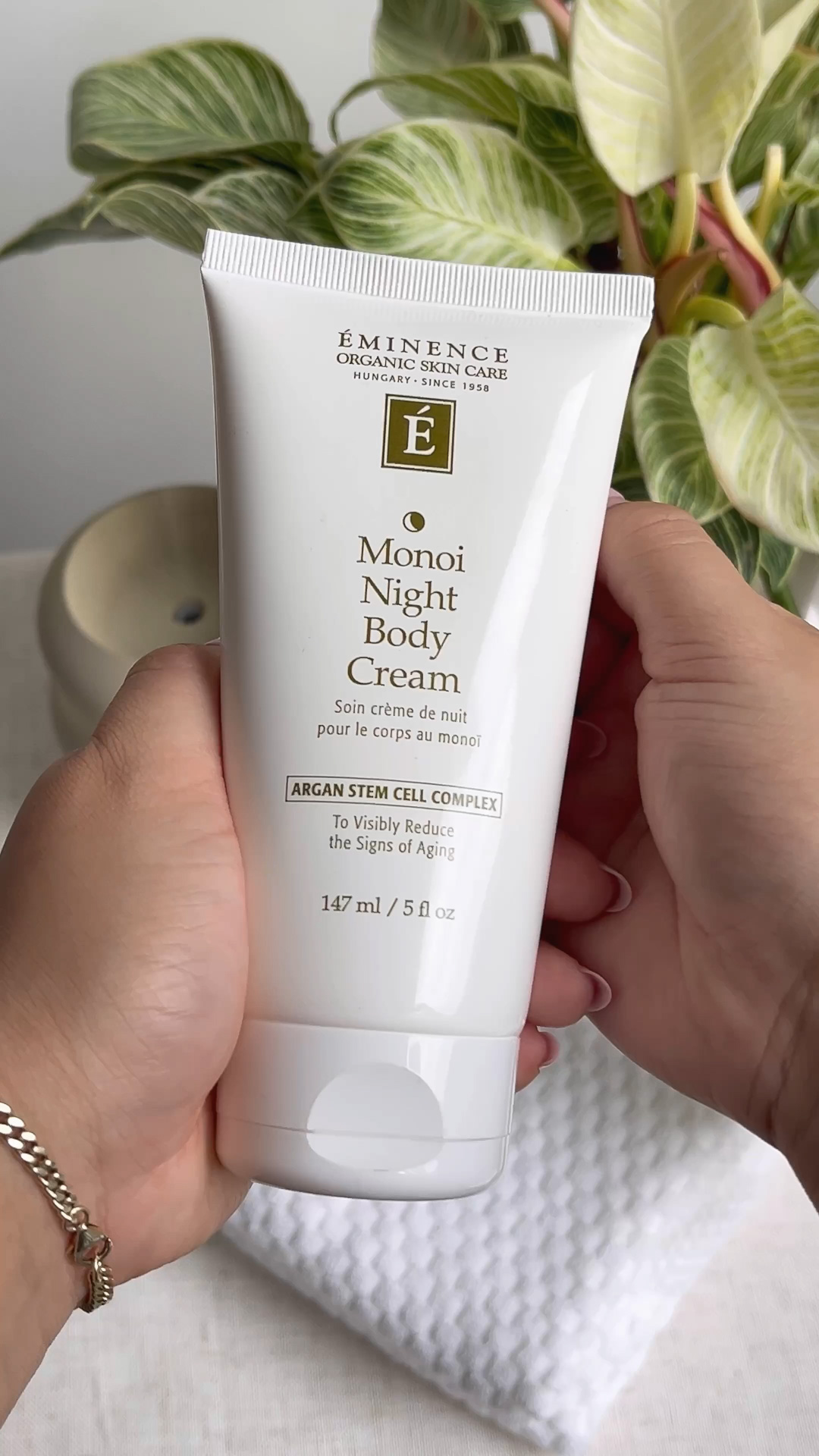 Two hands holding the Eminence Organics Monoi Age Corrective Night Body Cream tube