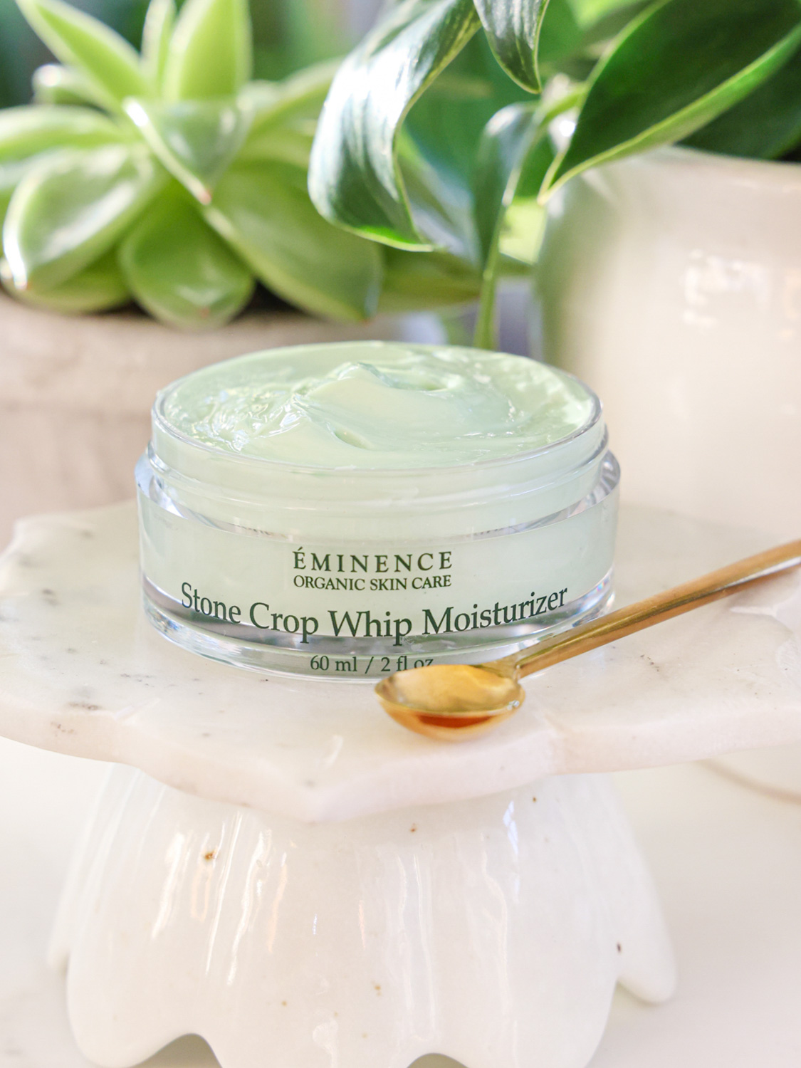 An open jar of the Eminence Organics Stone Crop Whip Moisturizer with a gold spoon on the side and with a background of green leaves