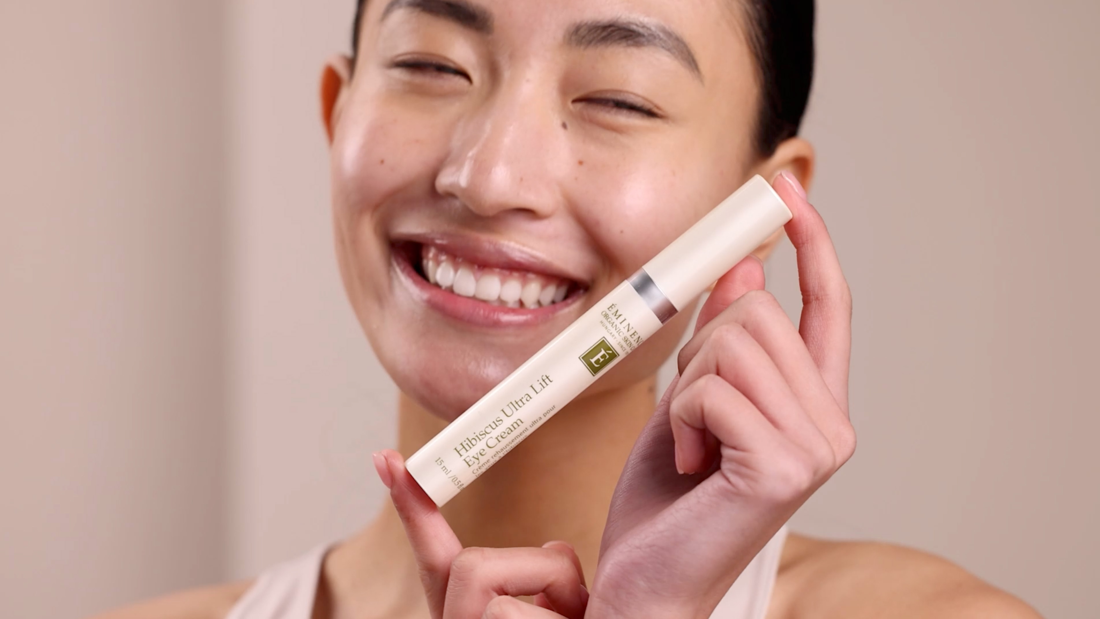 A woman holding the Eminence Organics Hibiscus Ultra Lift Eye Cream tube next to her face