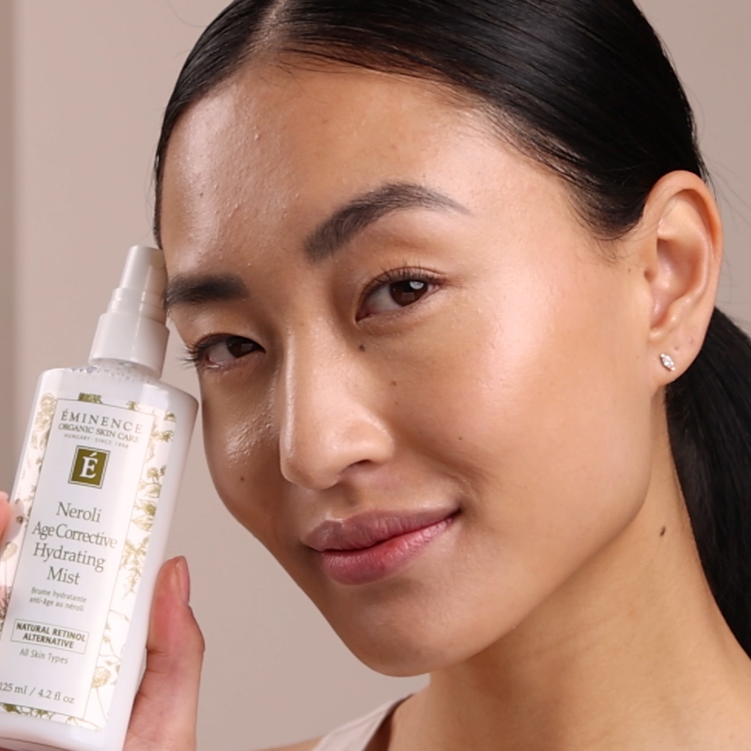 A woman holding the Eminence Organics Neroli Age Corrective Hydrating Mist bottle next to her face