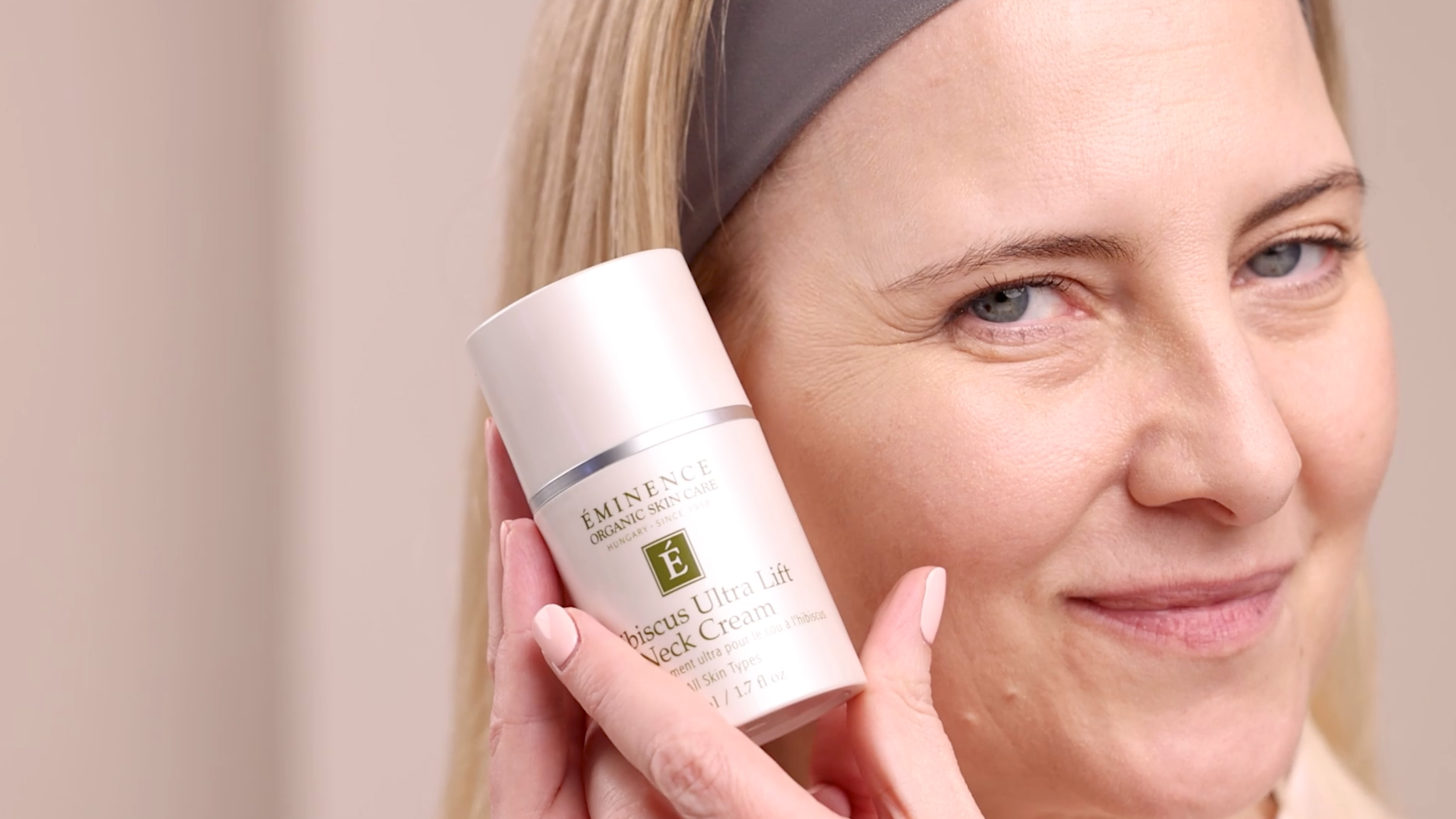 A woman holding the Eminence Organics Hibiscus Ultra Lift Neck Cream bottle next to her face