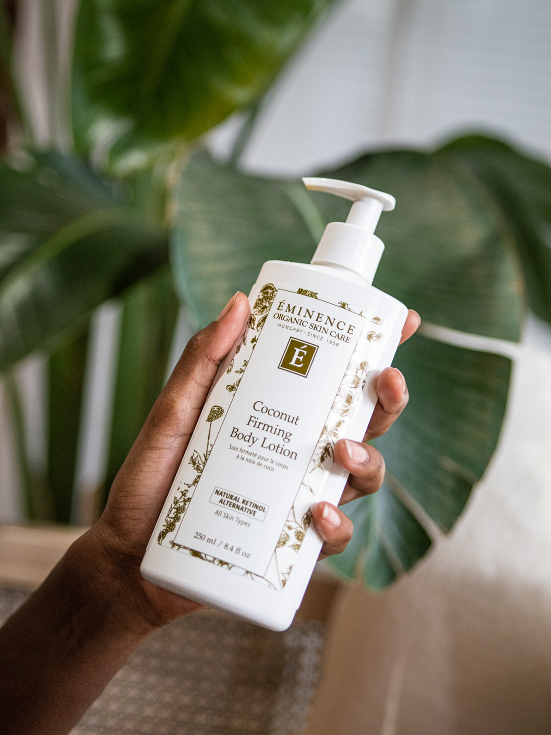 A hand holding the Eminence Organics Coconut Firming Body Lotion in front of a plant