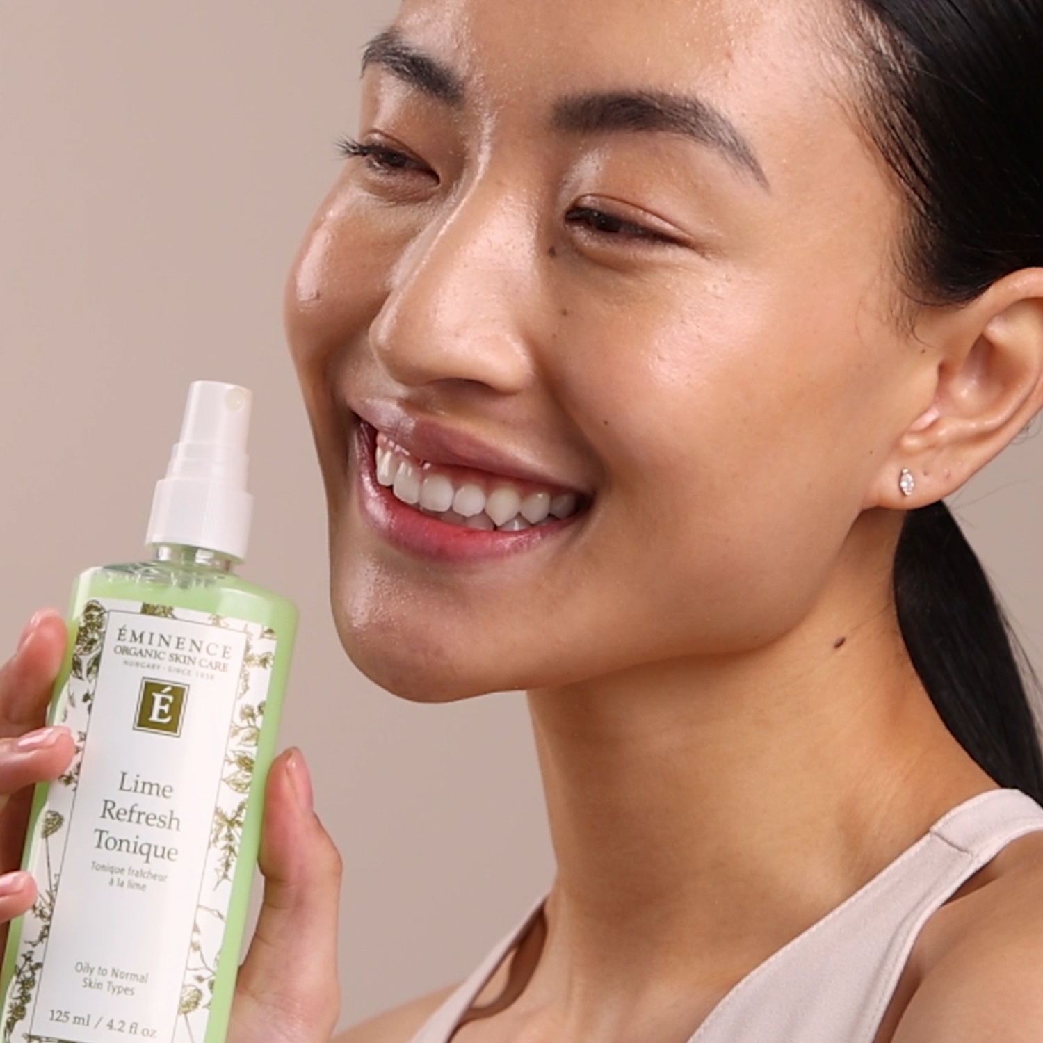 A woman holding the Eminence Organics Lime Refresh Tonique spray bottle next to her face