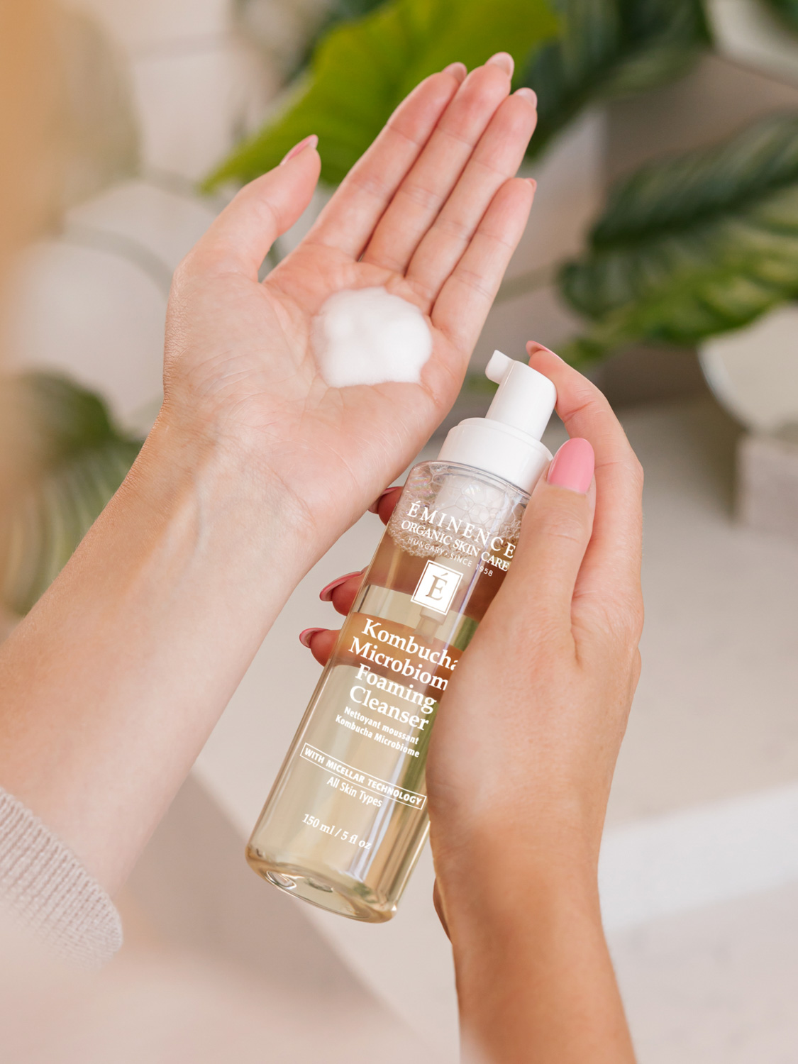 A woman holding the Eminence Organics Kombucha Microbiome Foaming Cleanser bottle in one hand and a swatch of the cleanser on the other hand