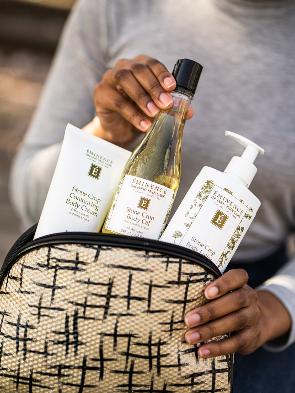 A hand grabbing the Eminence Organics Stone Crop Body Oil from a zippered pouch surrounded by other Eminence Organics Stone Crop products