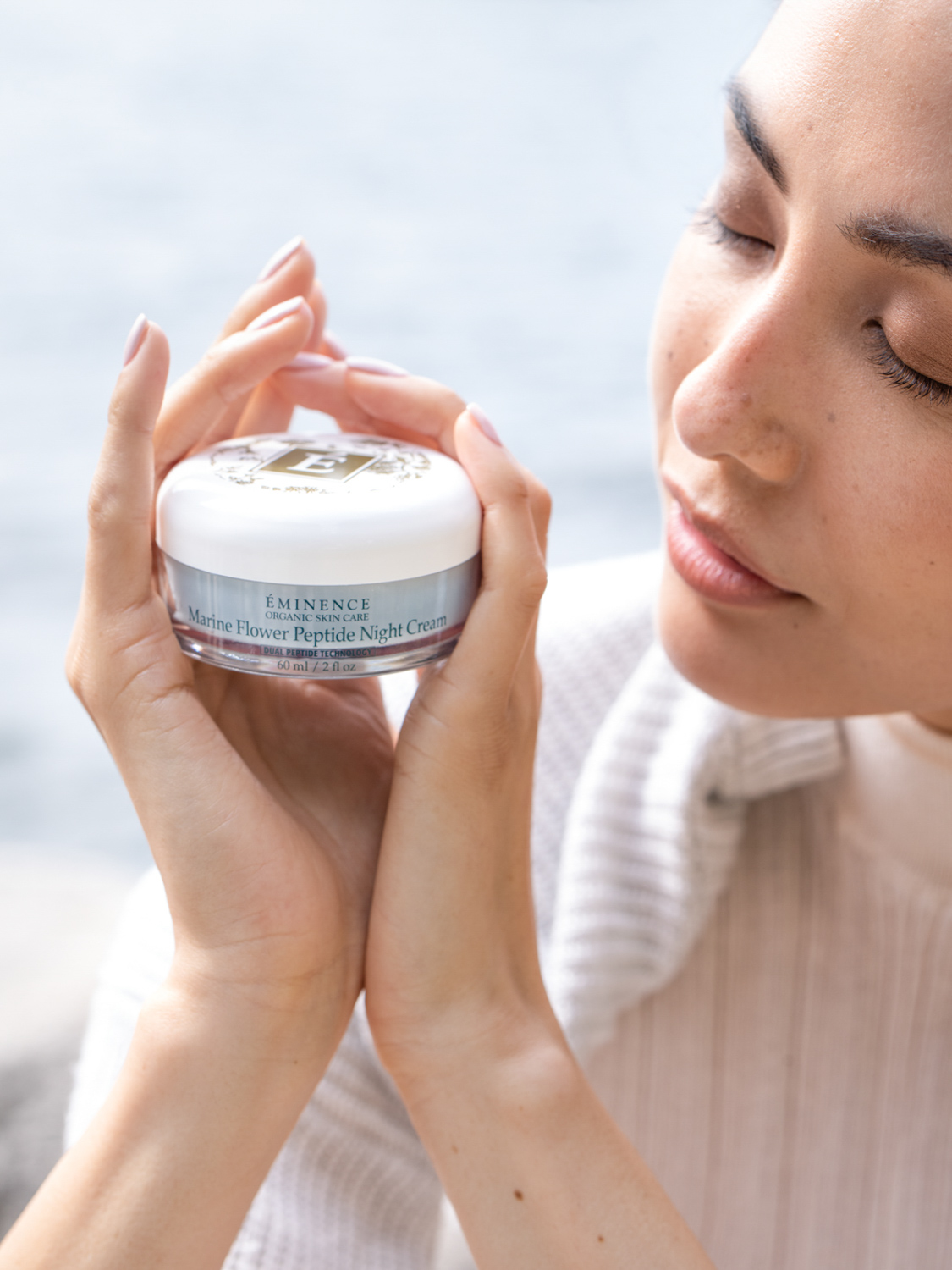 A woman holding the Eminence Organics Marine Flower Peptide Night Cream with both hands next to her face