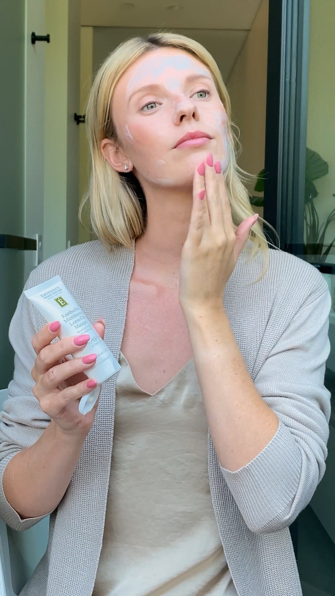 A woman applying the Eminence Organics Kombucha Microbiome Leave-On Masque on her face and holding the masque bottle with her other hand