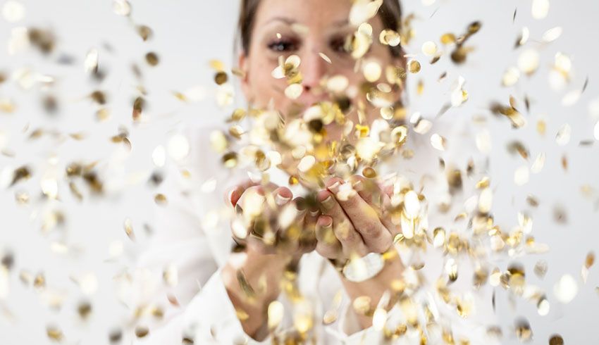 A woman' tosses gold confetti held in her cupped hands into the air, obscuring her face. 