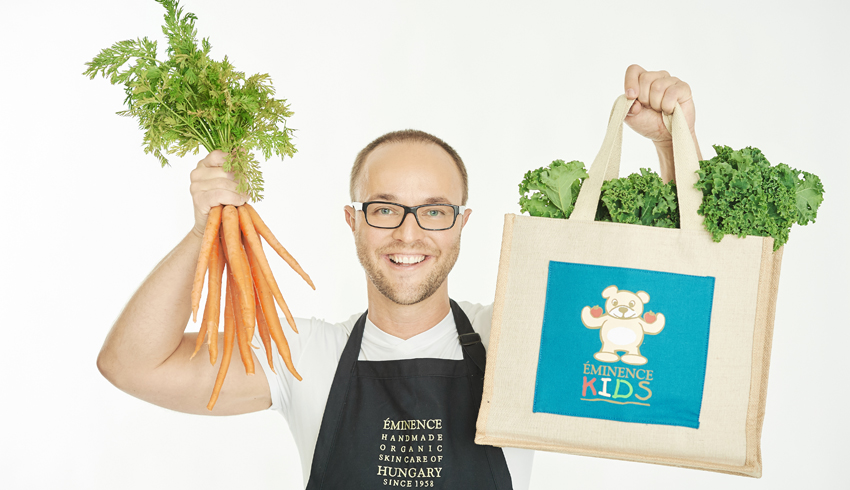 Éminence Organics President Boldijarre Koronczay holding up a bunch of fresh carrots in one hand and a grocery bag brimming with leafy greens in the other.