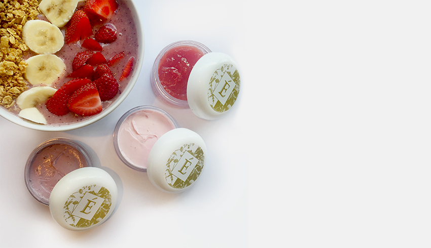 A smoothie bowl filled with granola, sliced bananas and sliced strawberries displayed next to three open containers of Eminence Organics Skin Care Masques. 