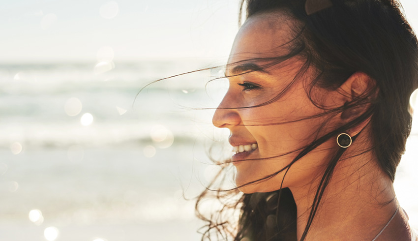 The profile of a smiling women whose hair is being blown into her face by the wind as she stands on the beach.. 