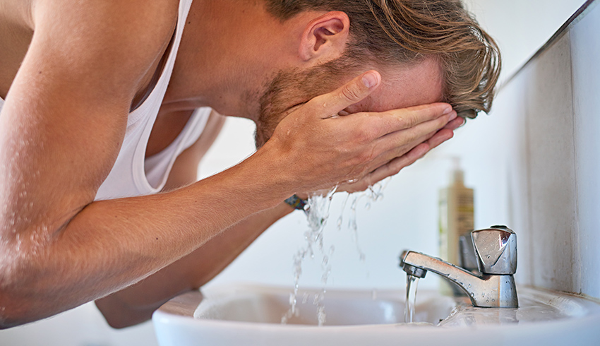 A man splashes water on his face as he bends over a sink with the faucet running. 