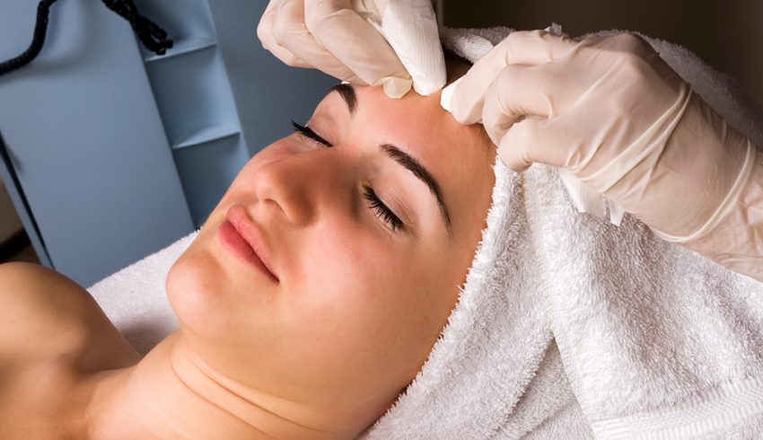 A woman with her hair wrapped in a towel lies back on a massage bed as the finger's of another woman's hands press down to extract dirt trapped in pores on area of her forehead. 