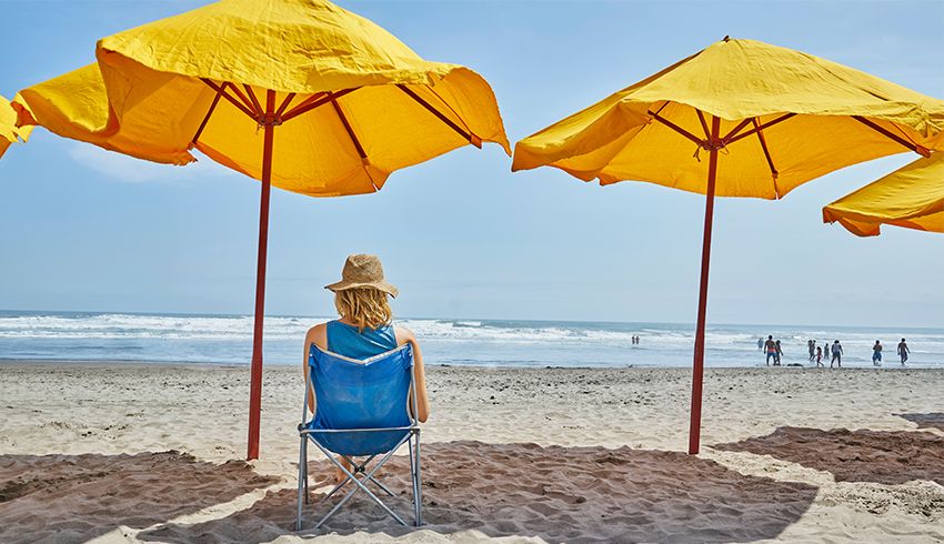 Woman sits in shade at the beach with a wide brimmed hat on.