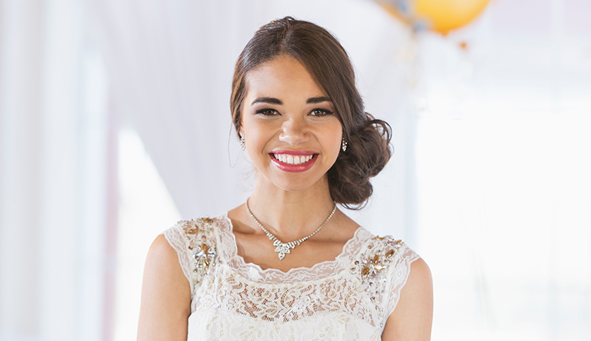 A young woman with her hair pinned to the side and wearing a white lace dress smiles. 