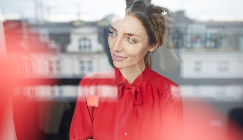 A woman in a red blouse is seen looking out a window. 