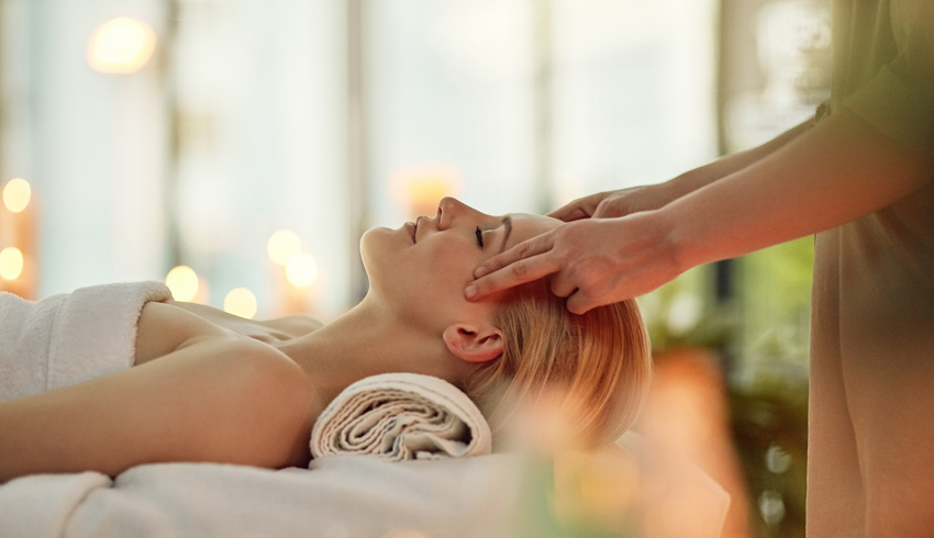 A woman with her eyes closed and a rolled towel supporting her neck lies on her back on a massage bed as another woman massages her temples. 