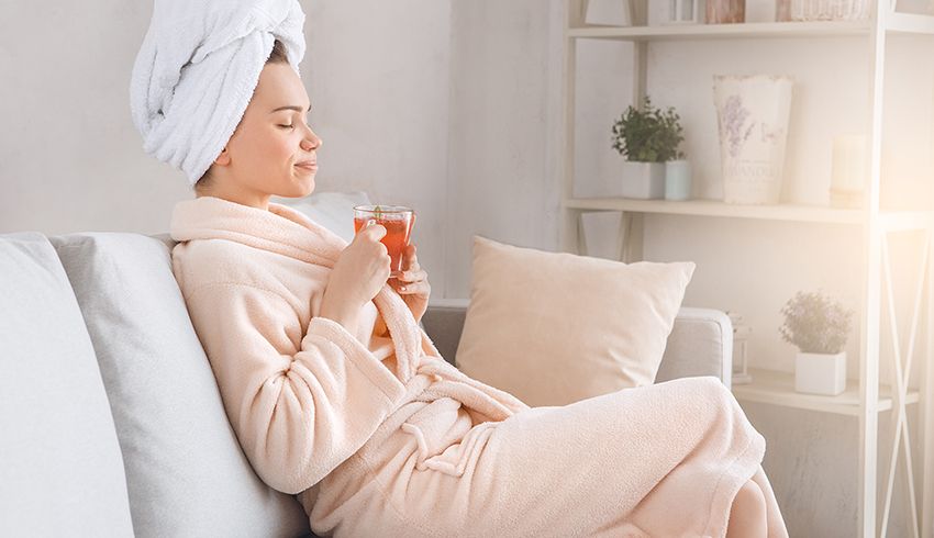 A woman wearing a bathrobe and a towel wrapped around her head sits back in a couch while holding a beverage in a clear glass mug. 