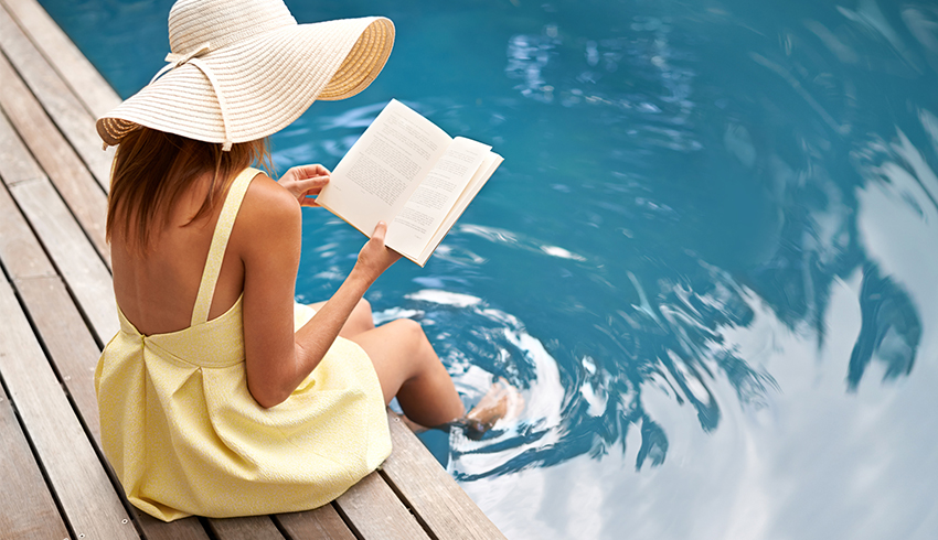 A woman wearing a yellow sundress and floppy star hat reads a book as she sits on the side of a dock with her legs dangling over the edge and her feet in the water. 