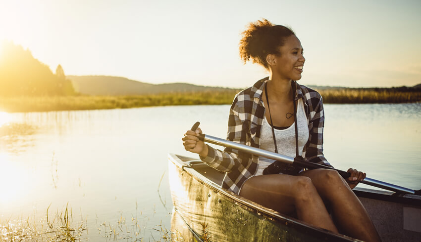 A smiling woman sits in a canoe on a lake as she holds a paddle in both hands. 