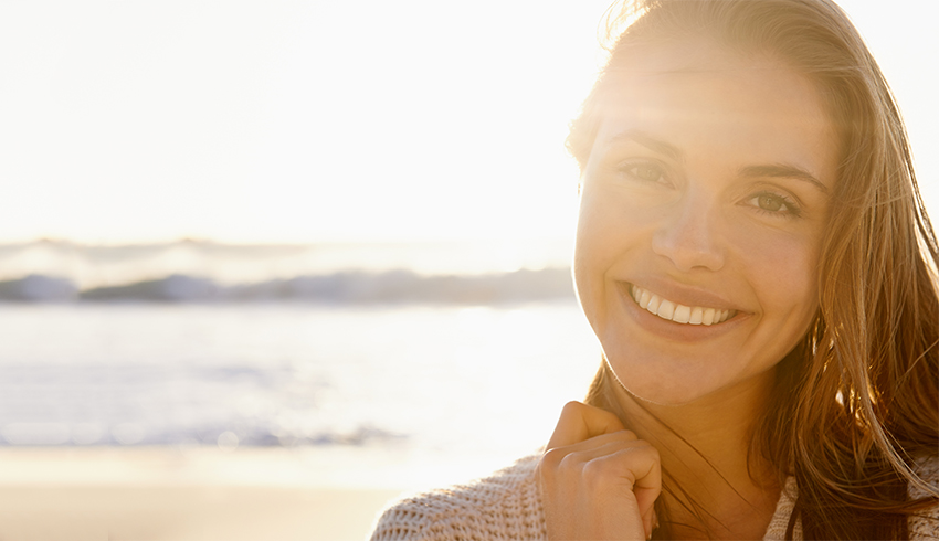 A smiling woman stands on a beach on a sunny day with the ocean in the background. 