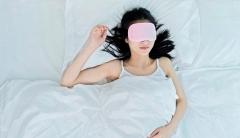 A woman wearing a pink sleep mask lies in a bed made of white satin sheets. 