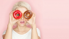 A woman holds two glazed donuts up to her eyes to imitate wearing a pair of glasses. 