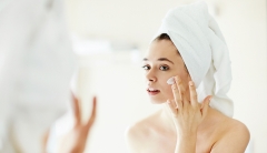 A woman with her hair wrapped in a towel looks at her reflection in a mirror as she applies lotion to her cheek. 