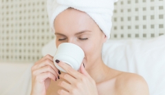 A woman with her hair wrapped in a towel sits in bed drinking from a mug. 
